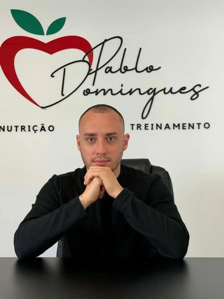 Pablo Domingues no consultório, em frente à parede com a logo Pablo Domingues — Nutrição e Treinamento